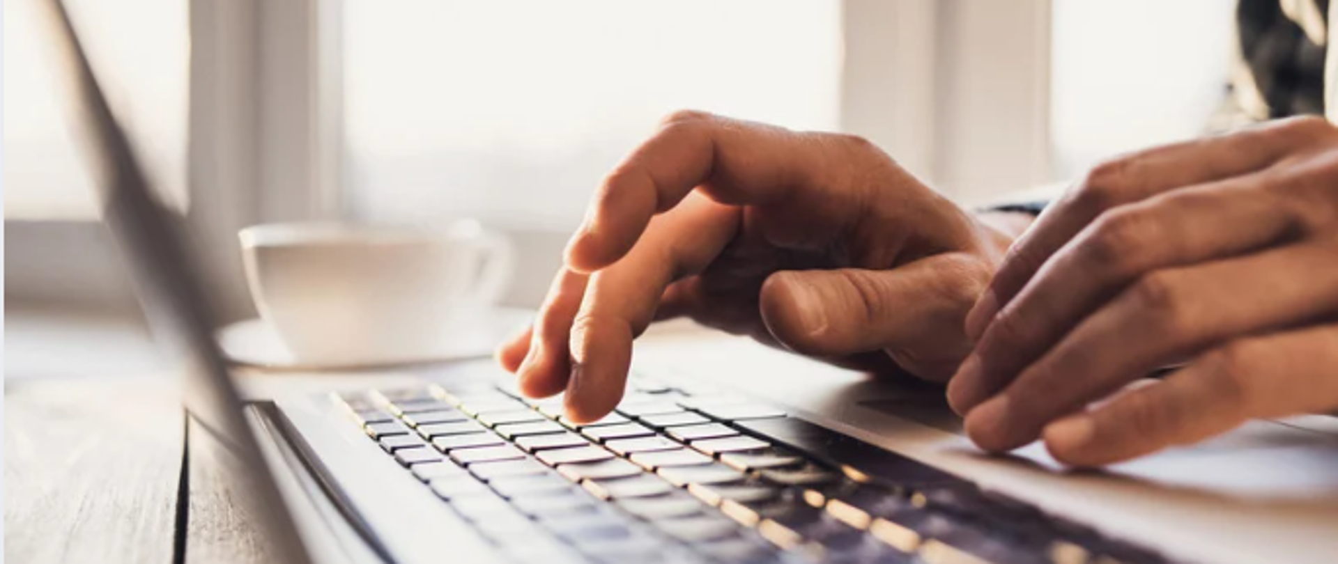 Zdjęcie przedstawia dłonie piszące na laptopie, przy laptopie stoi kawa