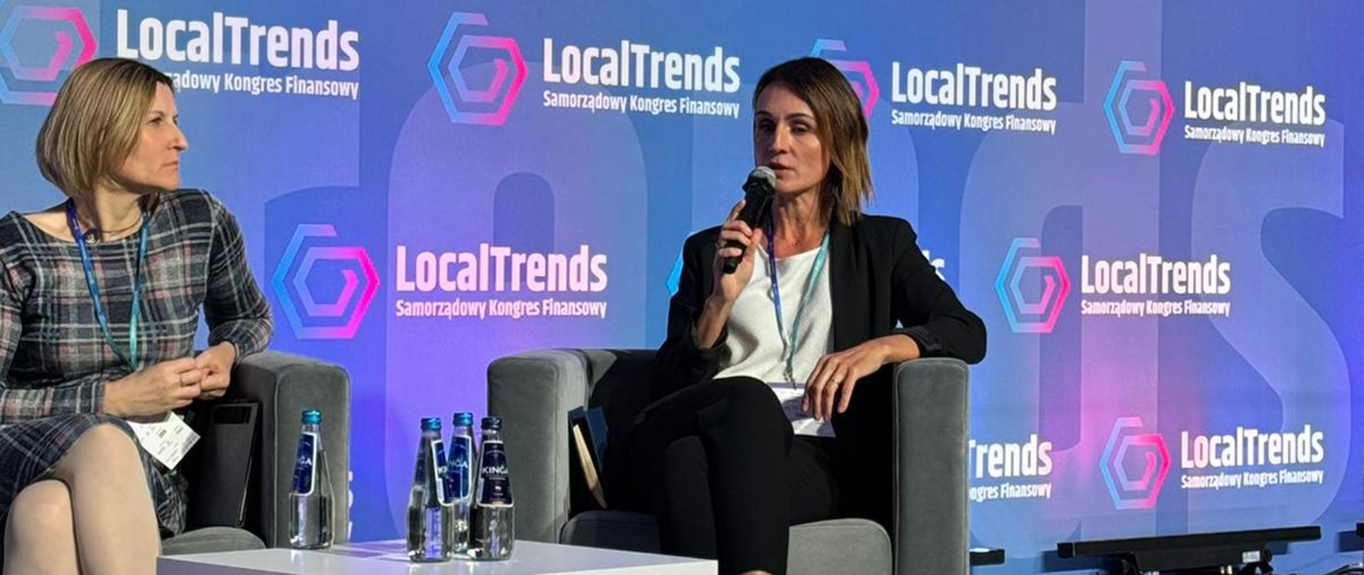 kobieta (wojewoda) siedzi na scenie i mówei do mikrofonu. w tle napis local trends