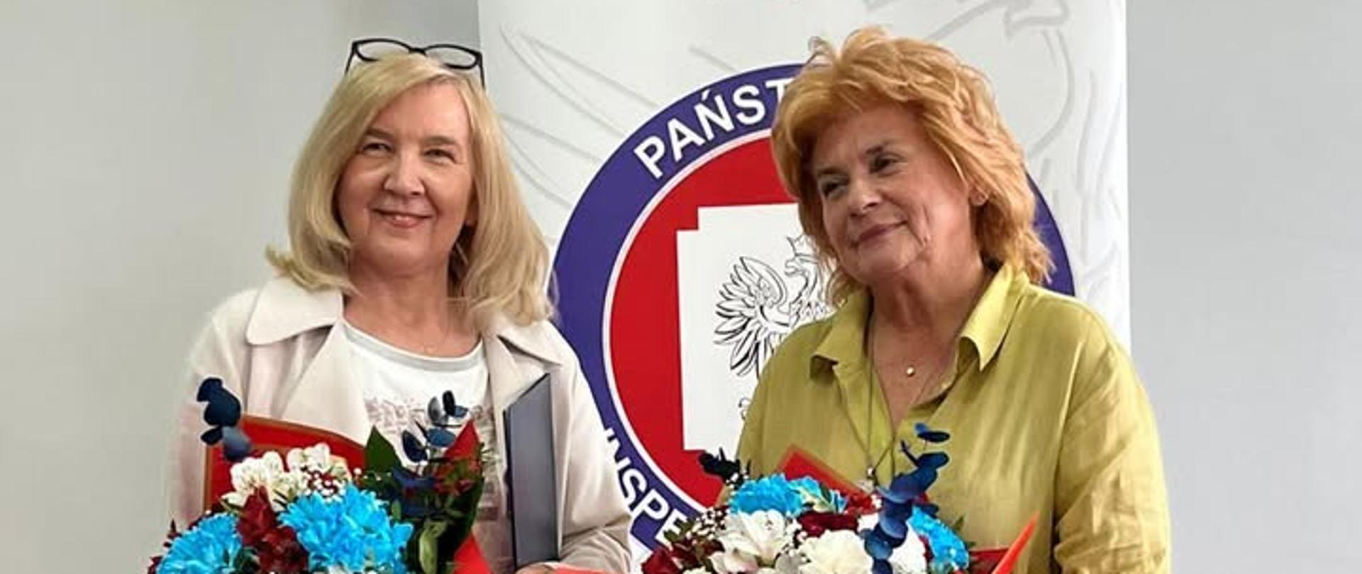 Na zdjęciu znajdują się dwie kobiety z kwiatami. Kobieta z prawej strony to Łódzki Wojewódzki Inspektor Sanitarny Urszula Jędrzejczyk z lewej powołana na Państwowego Powiatowego Inspektora Sanitarnego w Zgierzu Barbara Wieteska. Kobiety stoją na tle roll-upa z logo Inspekcji Sanitarnej