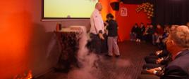 Mężczyzna w białej bluzie kucharskiej mówi do zgromadzonych. Wśród widzów również dzieci. Za nim na stoliku widoczny eksperyment w którym wydziela się słaniający o podłodze dym. 