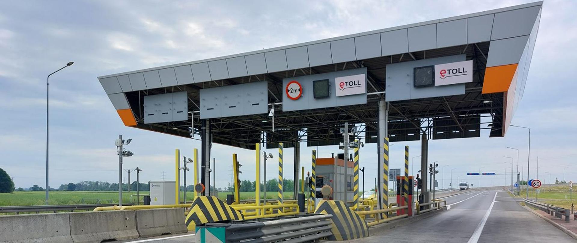Punkt poboru opłat na autostradzie A4 z zadaszonymi pasami przejazdowymi. Nad jezdnią znajdują się tablice z oznaczeniami systemu e-TOLL, kamery i urządzenia kontrolne. Po obu stronach pasów widoczne są bariery ochronne oraz słupy z czarno-żółtymi oznaczeniami. W tle rozciąga się płaski krajobraz z polami i pochmurnym niebem.