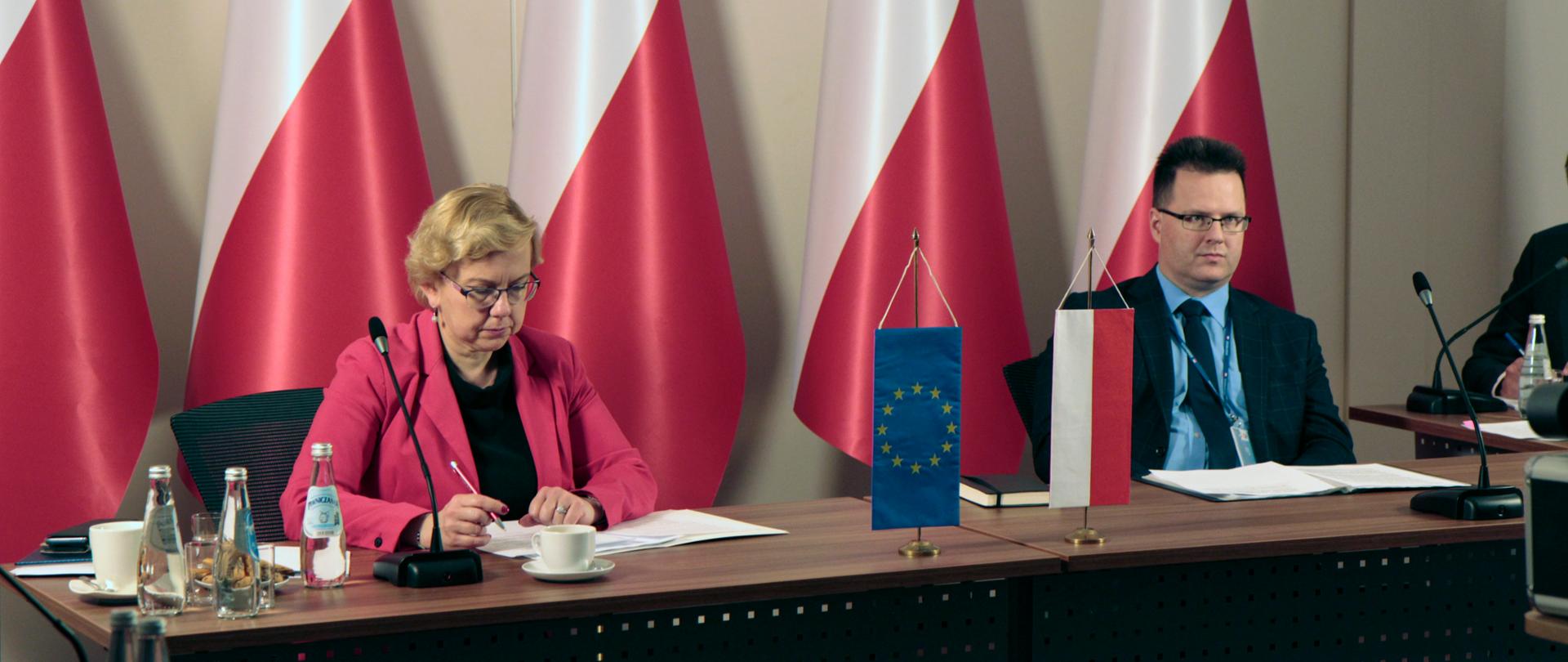 Na zdjęciu widać dwie osoby, które siedzą przy stole, są to kobieta i mężczyzna. Przed nimi leżą dokumenty, a w ich tle widać biało-czerwone flagi. Na środku stołu stoi mała flaga, symbol Unii Europejskiej