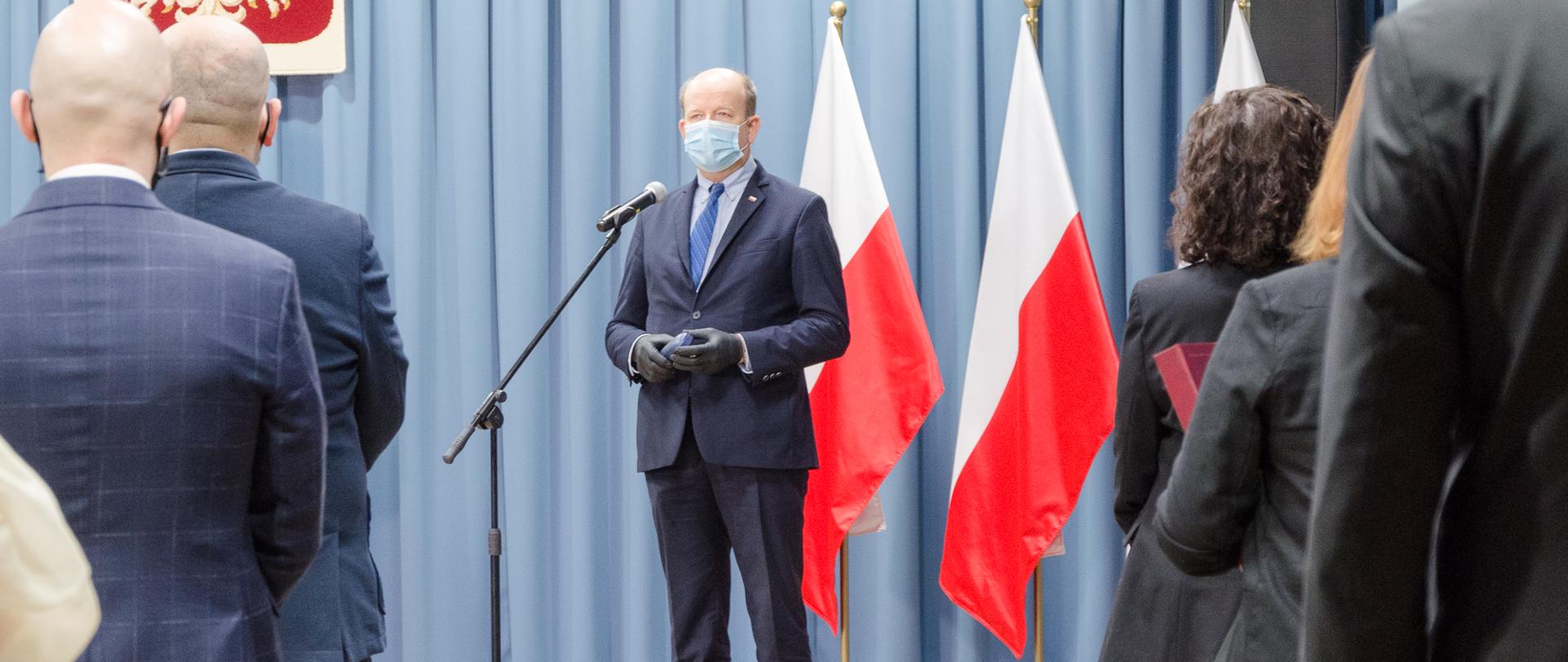 Bo bokach zdjęć znajdują się elegancko ubrani ludzie (operatorzy numeru alarmowego 112). Po środku znajduje się elegancko ubrany mężczyzna, który przemawia na tle biało czerwonych flag