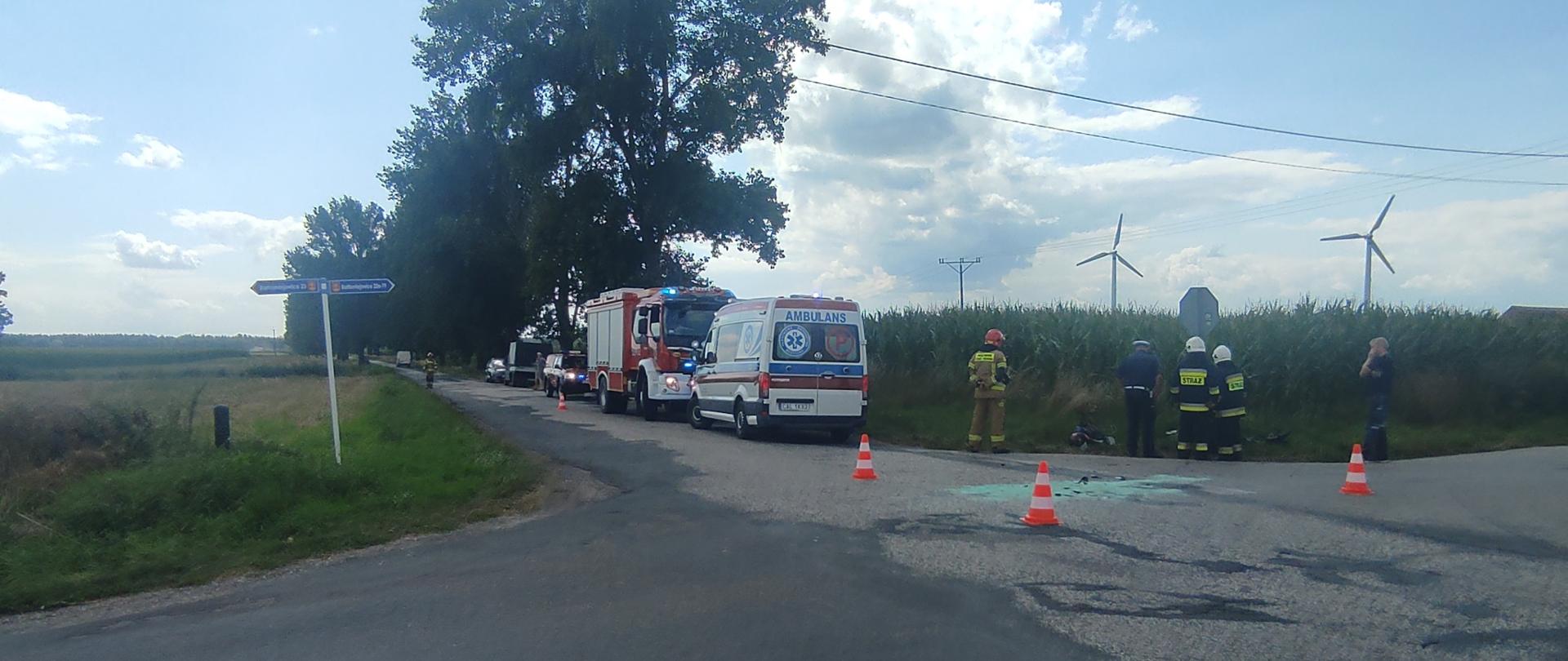 Zdjęcie przedstawia skrzyżowanie drogi wojewódzkiej oraz powiatowej, na której znajdują się pojazdy służb ratowniczych (dwa wozy straży pożarnej, pogotowie ratunkowe) strażaków i policjantów podczas czynności po kolizji drogowej pojazdu osobowego z motocyklem