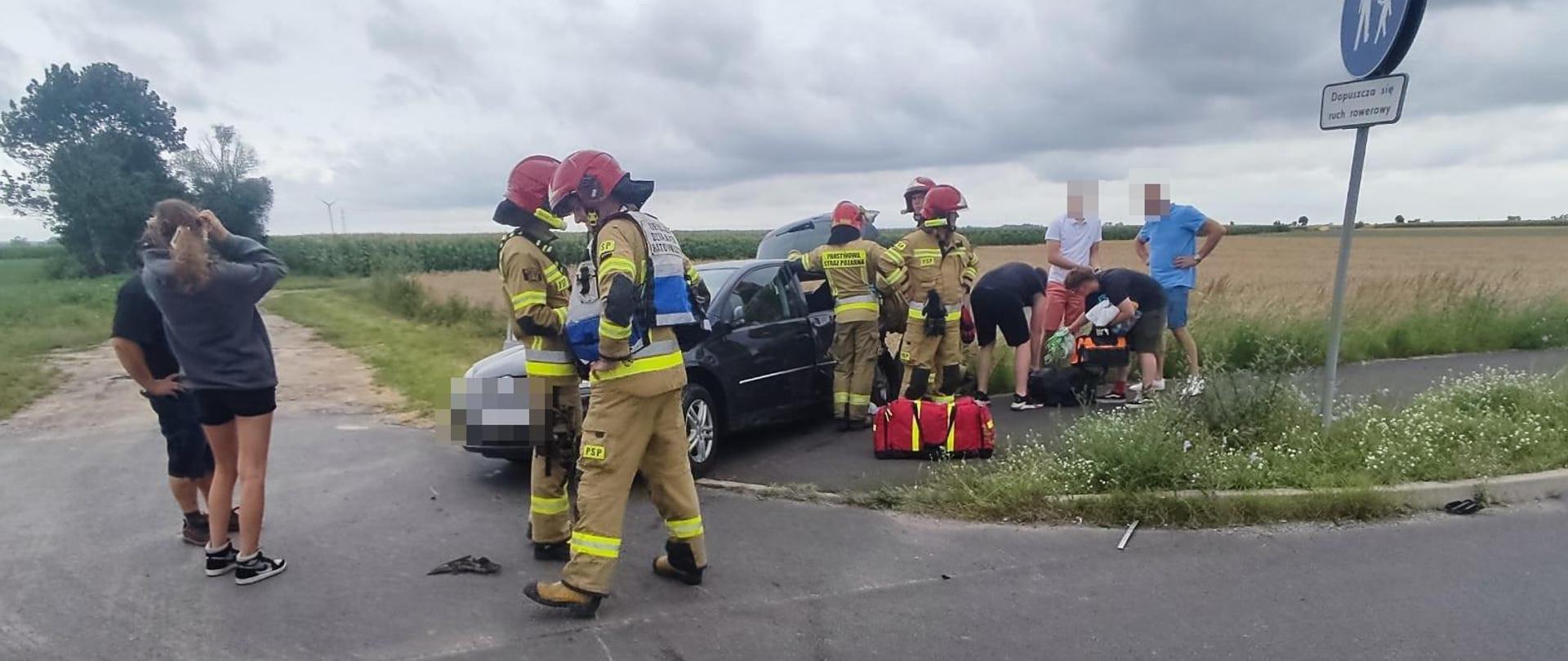 Wypadek na skrzyżowaniu dróg. Widoczny pojazd z rozbitym tyłem. Obok pojazdu strażacy udzielają pomocy medycznej uczestnikom zdarzenia. 