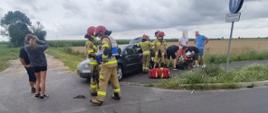 Wypadek na skrzyżowaniu dróg. Widoczny pojazd z rozbitym tyłem. Obok pojazdu strażacy udzielają pomocy medycznej uczestnikom zdarzenia. 