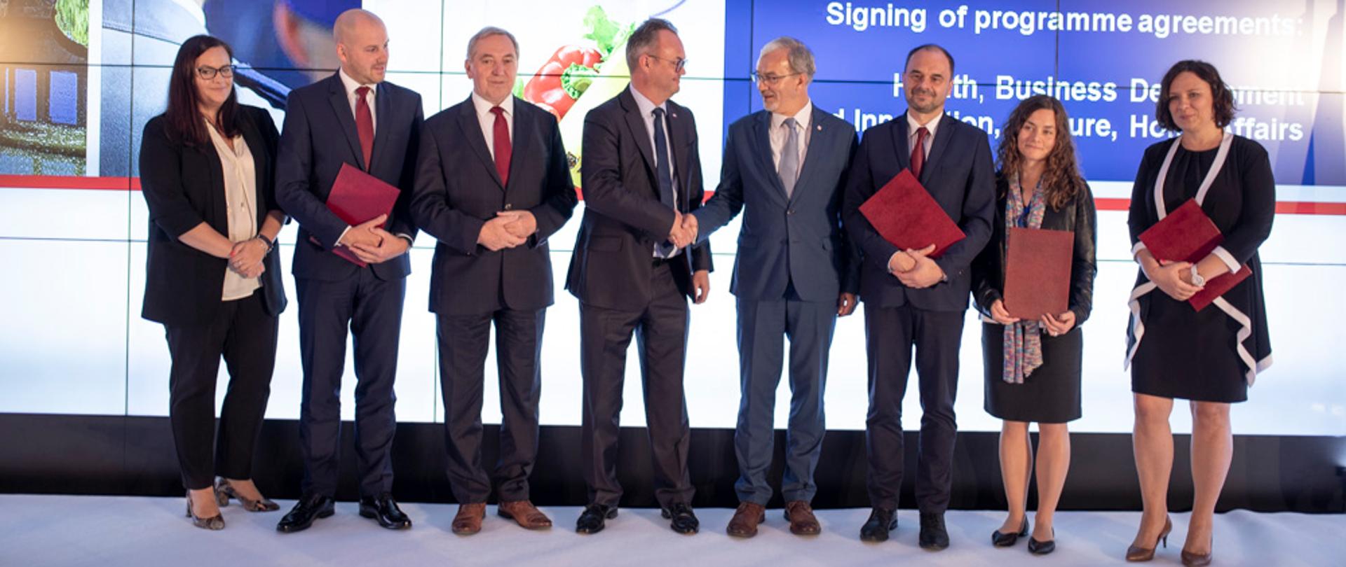 Osiem osób na scenie, w środku ambasador i minister podają sobie dłonie, w tle ekran z napisem "Podpisanie umów ws. programów: Zdrowie, Rozwój Przedsiębiorczości i Innowacje, Kultura, Sprawy Wewnętrzne"