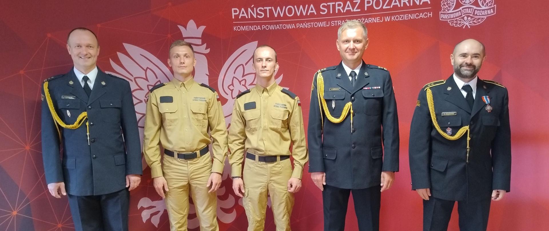 funkcjonariusze w ubraniach służbowych stoją na tle ścianki w świetlicy KP PSP Kozienice