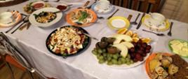 Świąteczny stół Wielkanocny przykryty białym obrusem, na którym znajdują się kolorowe talerze z potrawami, ciastem i owocami.