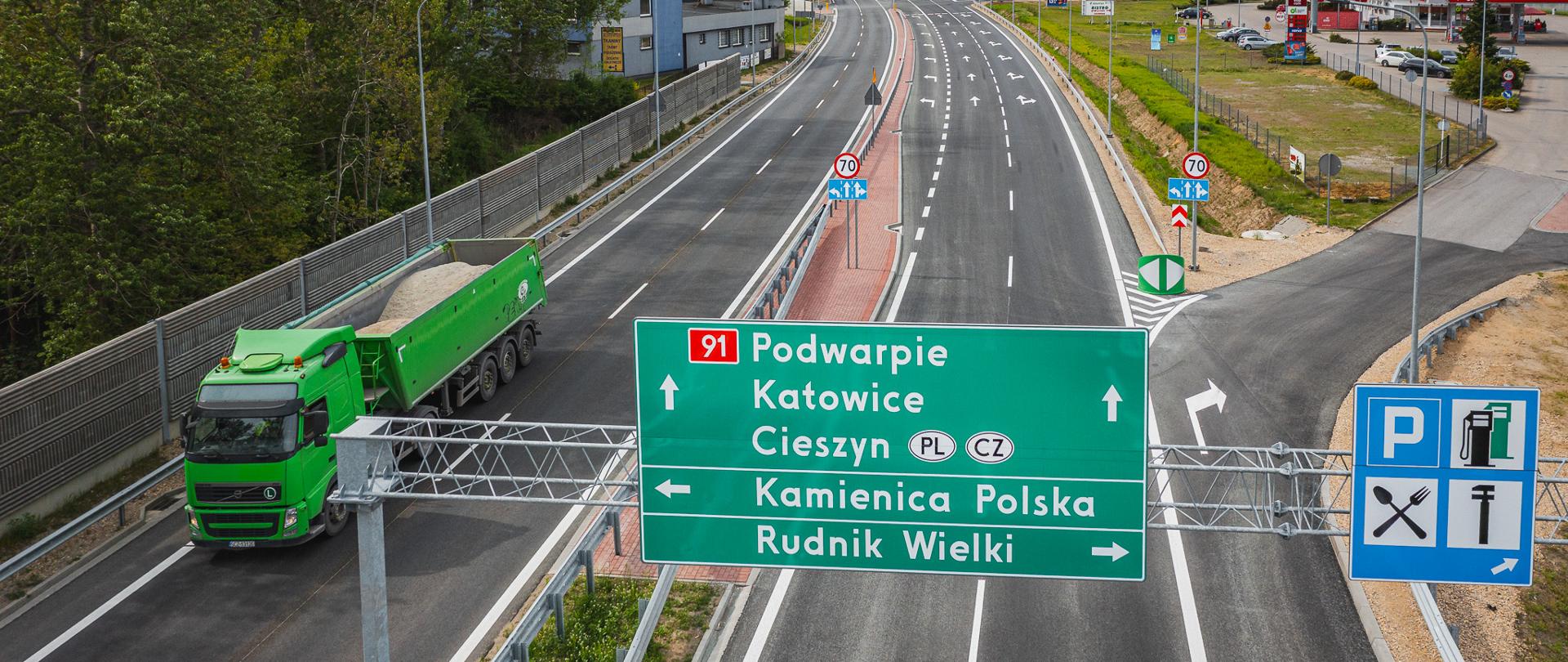 Odcinek drogi krajowej nr 91 w kierunku Podwarpia, Katowic i Cieszyna, z widocznymi znakami kierunkowymi, zatoką zjazdową na Kamienicę Polską i Rudnik Wielki oraz pojazdami ciężarowymi.