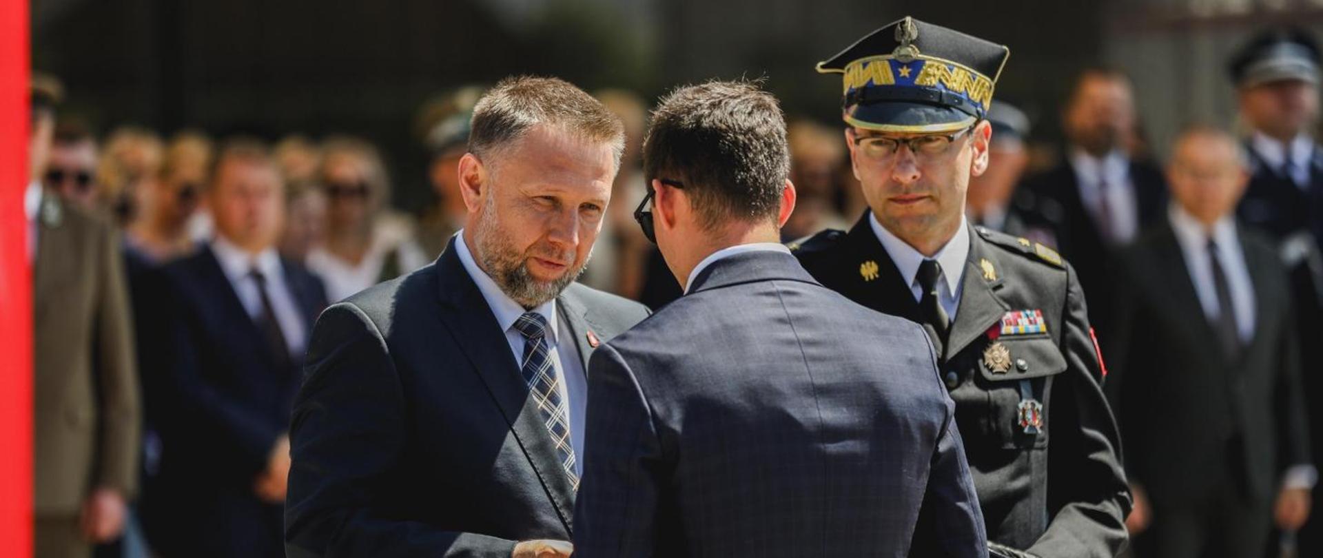 Mężczyzna w garniturze wręcza wyróżnienie uczestnikowi uroczystości podczas obchodów strażackich na placu apelowym. Obok stoi komendant główny PSP w galowym mundurze, a w tle widoczni są zaproszeni goście i funkcjonariusze uczestniczący w wydarzeniu.