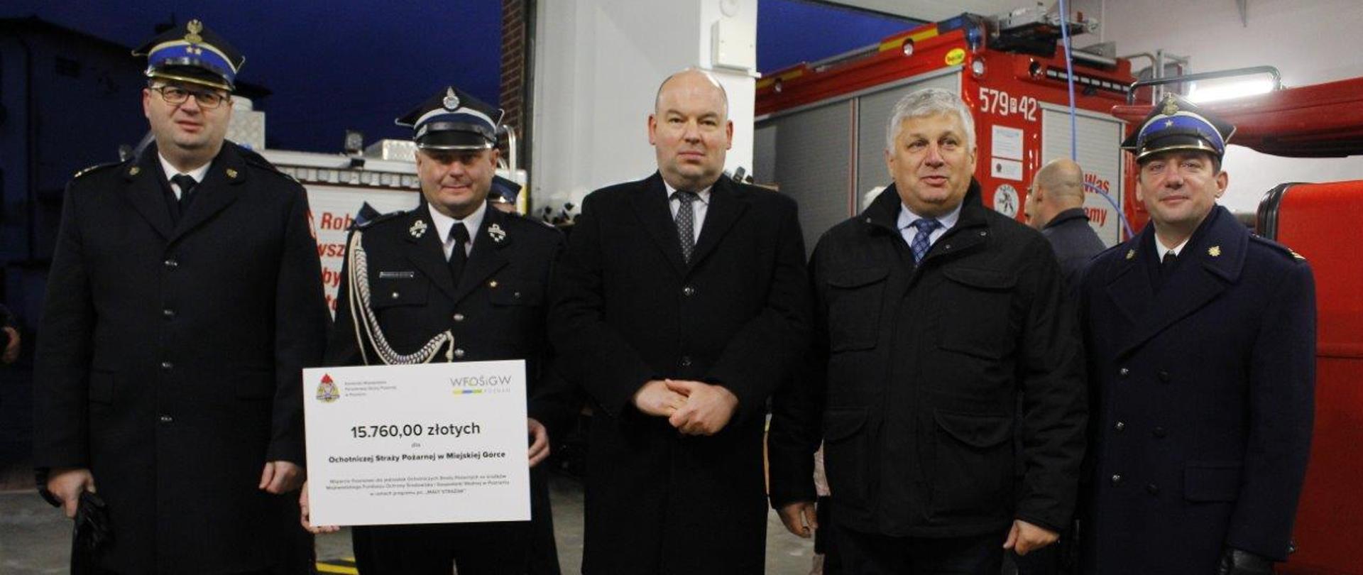 grupa strażaków oraz osób cywilnmych stoi przed pojazdem strażackim, jedna osoba trzyma dużą białą kartkę z kwotą dofinasnowania