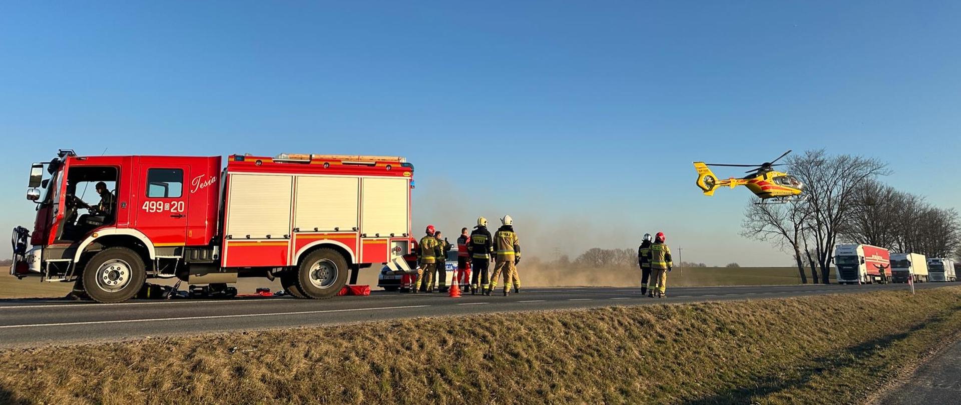 Na zdjęciu znajduje się wóz bojowy,strażacy startujący śmigłowiec a w tle samochody ciężarowe