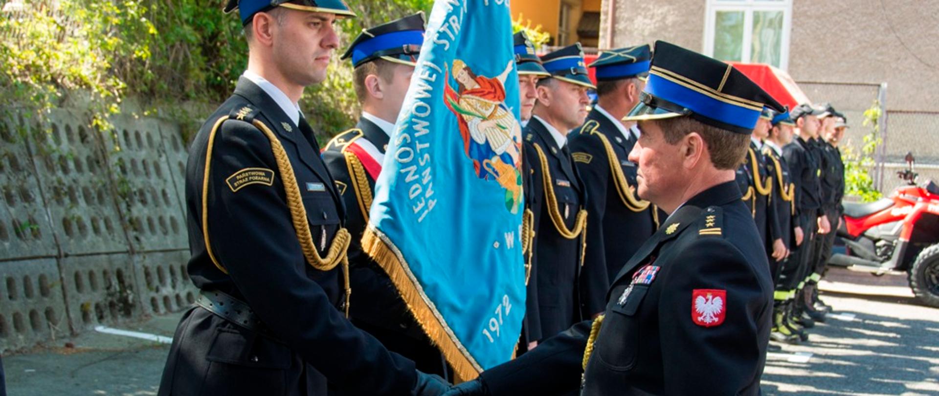 Zdjęcie wykonane na zewnątrz. Na zdjęciu strażacy Państwowej Straży Pożarnej ubrani w mundury galowe, ustawieni są w szeregu na uroczystej zbiórce. Na pierwszym planie strażak Państwowej Straży Pożarnej w stopniu starszego brygadiera podaje dłoń strażakowi w stopniu starszego kapitana.