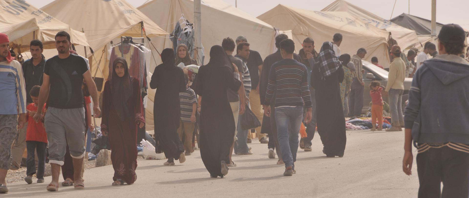 Ulica w obozie dla uchodźców, po której idą w różnych kierunkach kobiety w strojach muzułmańskich, dzieci i mężczyźni. Wzdłuż ulicy ustawione są namioty w kolorze beżowym.