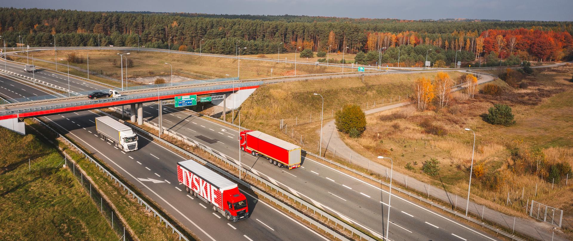 Zdjęcie przedstawia autostradę z lotu ptaka, na której znajdują się trzy ciężarówki. Dwie z nich jadą w jednym kierunku, a jedna w przeciwnym. Na pierwszym planie widoczna jest ciężarówka z napisem "TYSKIE" na naczepie. W tle widać wiadukt, przez który przejeżdża samochód osobowy. Wokół autostrady rozciągają się tereny zielone z drzewami, które mają jesienne barwy. Na wiadukcie znajdują się znaki drogowe, które wskazują kierunki jazdy. Zdjęcie jest interesujące ze względu na perspektywę z lotu ptaka, która pozwala zobaczyć układ dróg oraz otaczający krajobraz. 