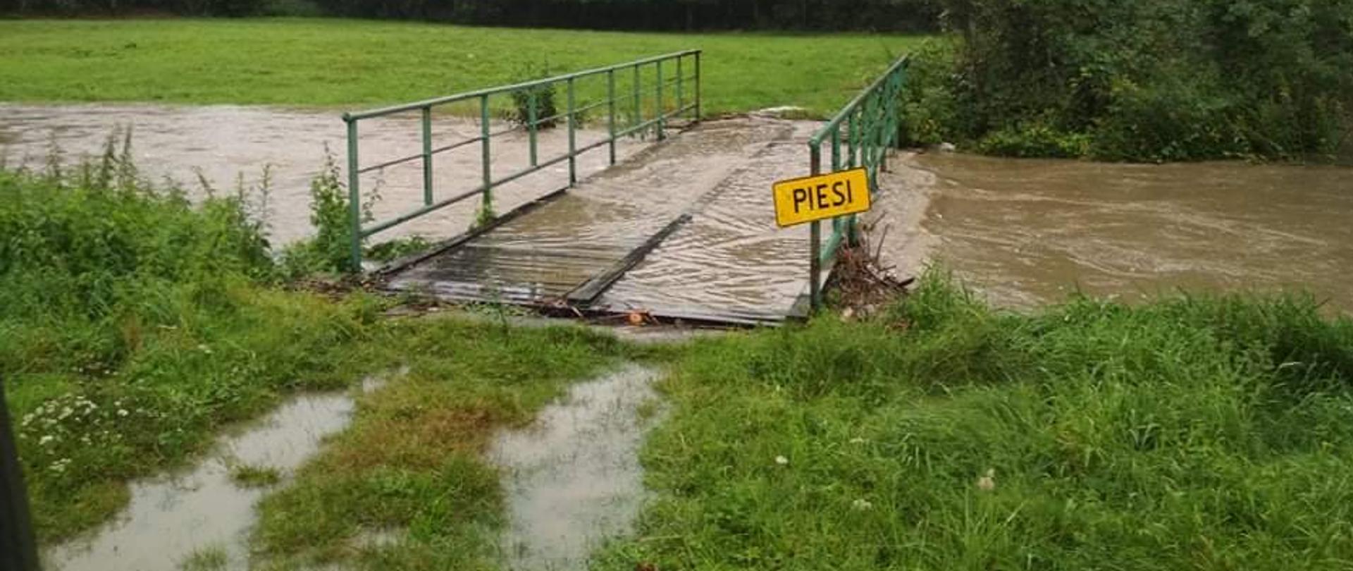 Na zdjęciu widoczny potok Kisielina w znacznym stopniu podniesionym poziomem wody nad potokiem znajduje się kładka dla pieszych przez którą przelewa się woda. Z prawej strony widoczny napis koloru czarnego na tle żółtym „Piesi’. 
