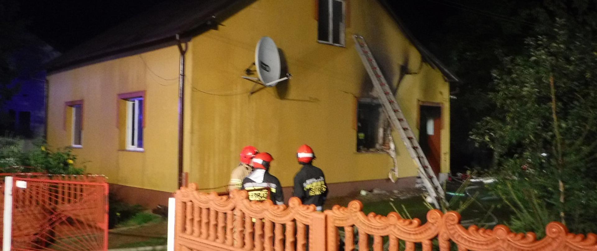 Na zdjęciu widzimy trzech strażaków w pełnym uzbrojeniu. Widzimy cześć ogrodzenia betonowego a w głębi budynek mieszkalny z wybitym od wybuchu oknem, o ścianę budynku oparta jest drabina.