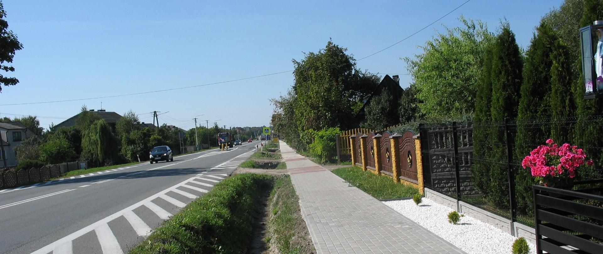 Droga krajowa jednojezdniowa, wzdłuż drogi rów, chodnik, ogrodzenia posesji. drogą jadą samochody osobowe i ciężarowe