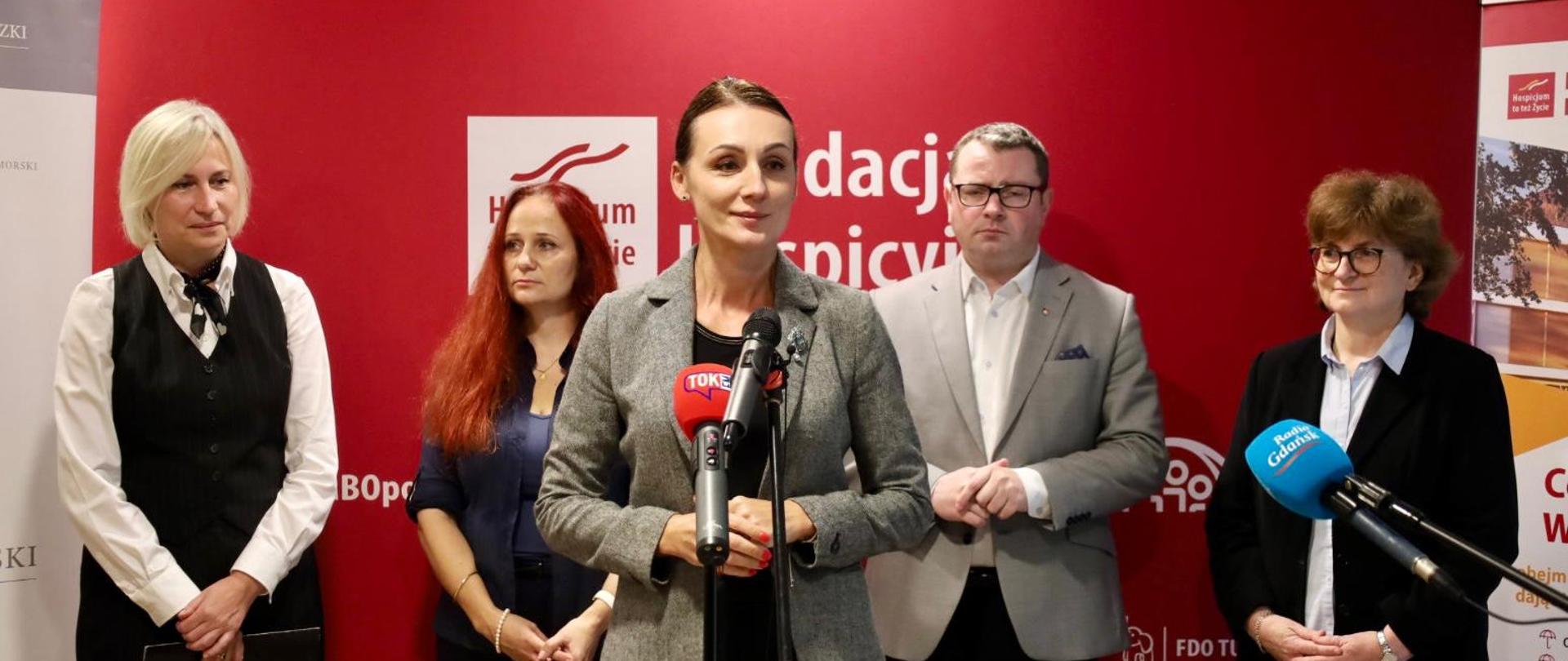 Na zdjęciu jest pięć osób. Z tyłu stoją cztery - trzy kobiety i mężczyzna. Na pierwszym planie, przy mikrofonach, stoi kobieta. W tle czerwona ścianka z napisem "Fundacja Hospicyjna".