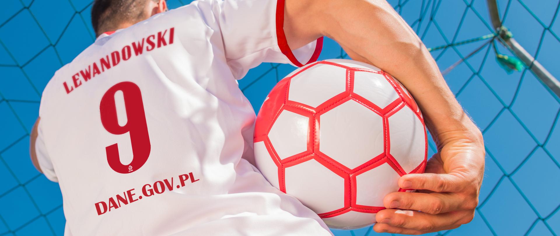 Zdjęcie pleców chłopaka w biało-czerwonej piłkarskiej koszulce, stoi na tle bramki, w prawym ręku trzyma piłkę. Na koszulce napis: Lewandowski, numer 9 i adres dane.gov.pl