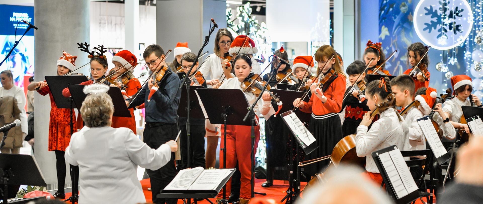 Kolorowe zdjęcie zespołu instrumentalnego w skład którego wchodzą dzieci grające na skrzypcach, wiolonczelach i kontrabasie. Dzieci stoją na scenie i grają na instrumentach, ubrane są w eleganckie stroje w kolorach czerwonych, czarnych i białych oraz elementy świąteczne typu czapki mikołaja, rogi renifera. Przed zespołem stoi Pani dyrygent, która ma na głowie aureolę, przed nią znajduje się pulpit z nutami.