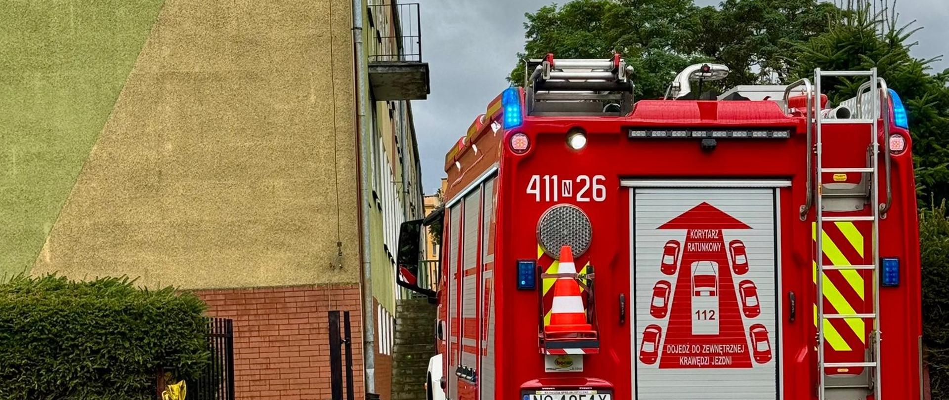 Duży czerwony samochód strażacki stoi we wjeździe na teren szkoły. Z lewej strony budynek szkoły.