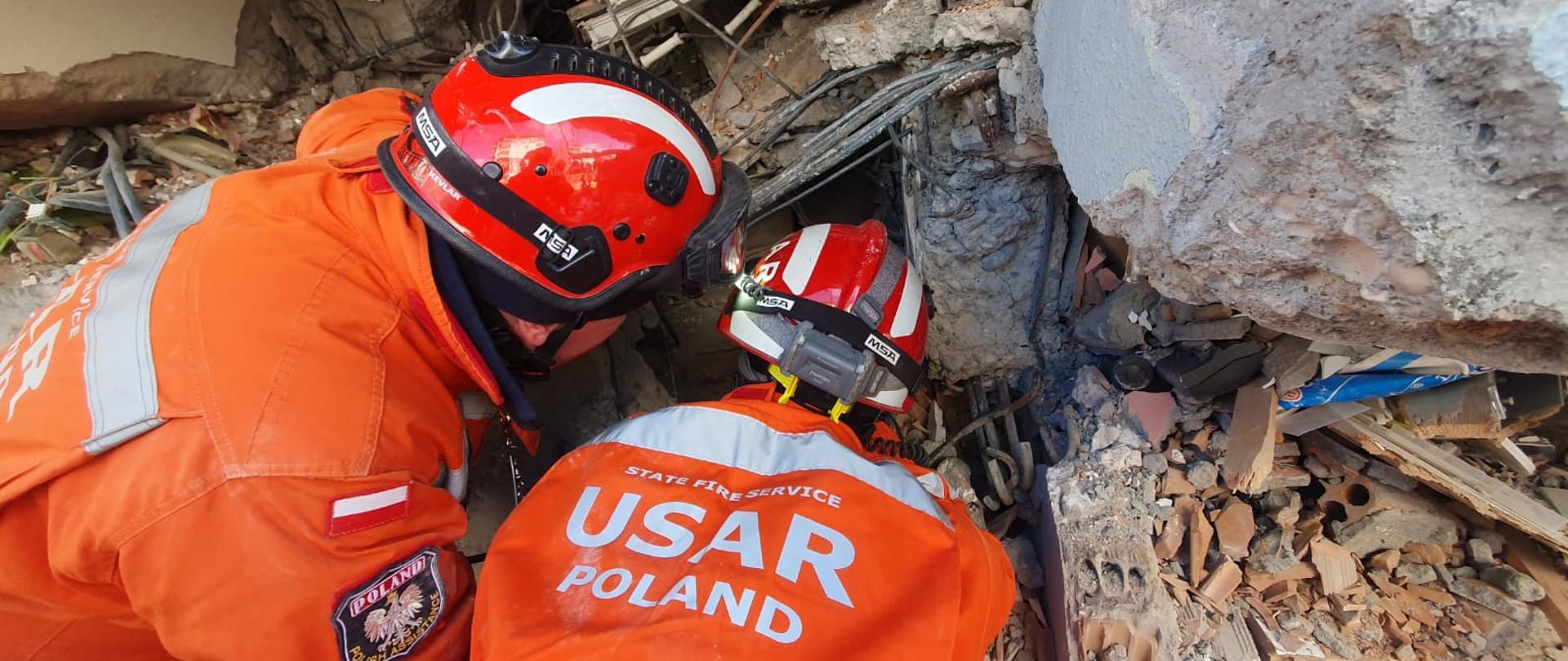 Dwóch ratowników strażaków z napisem na umundurowaniu USAR POLAND zagląda w szczelinę na gruzowisku