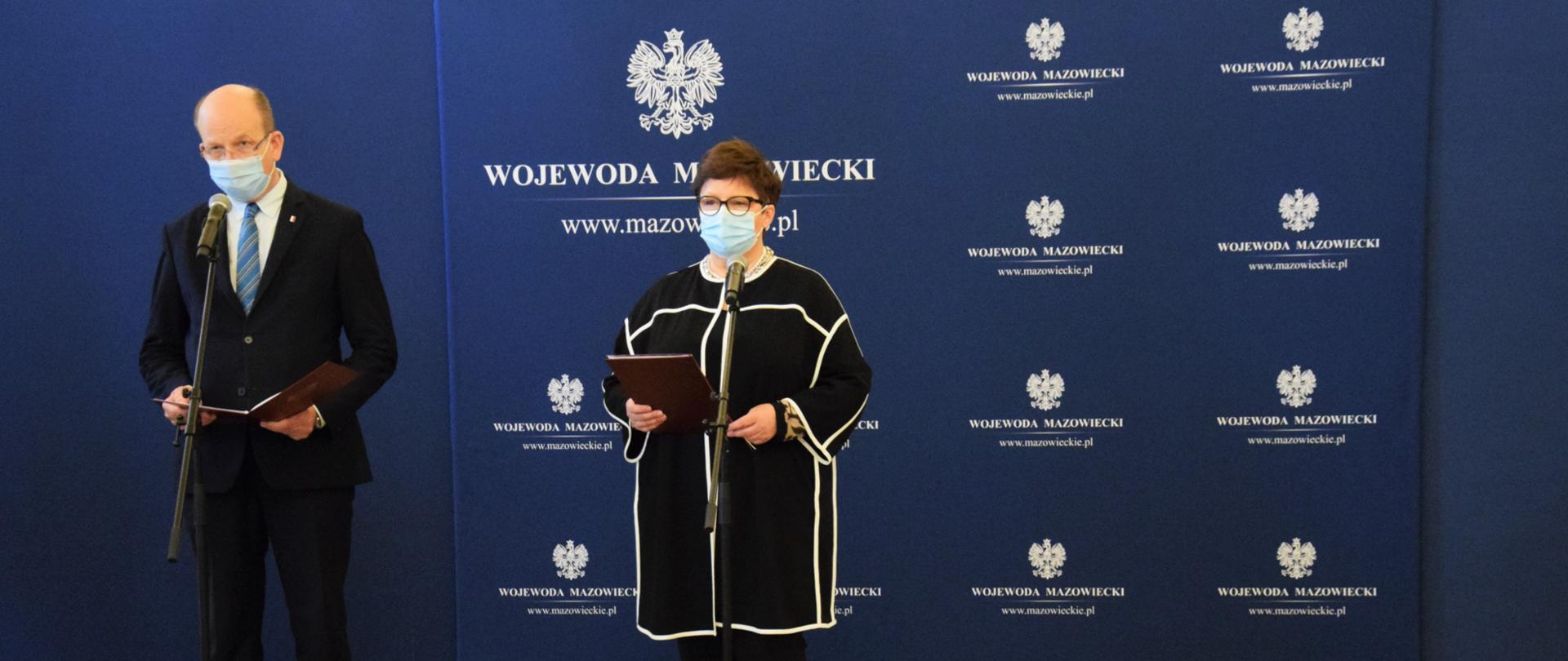 Od lewej: Wojewoda Mazowiecki ubrany w ciemny garnitur z niebieskim krawatem, stojący przy mikrofonie, obok
Ewa Więckowska ubrana w ciemny strój, stojąca przy mikrofonie.