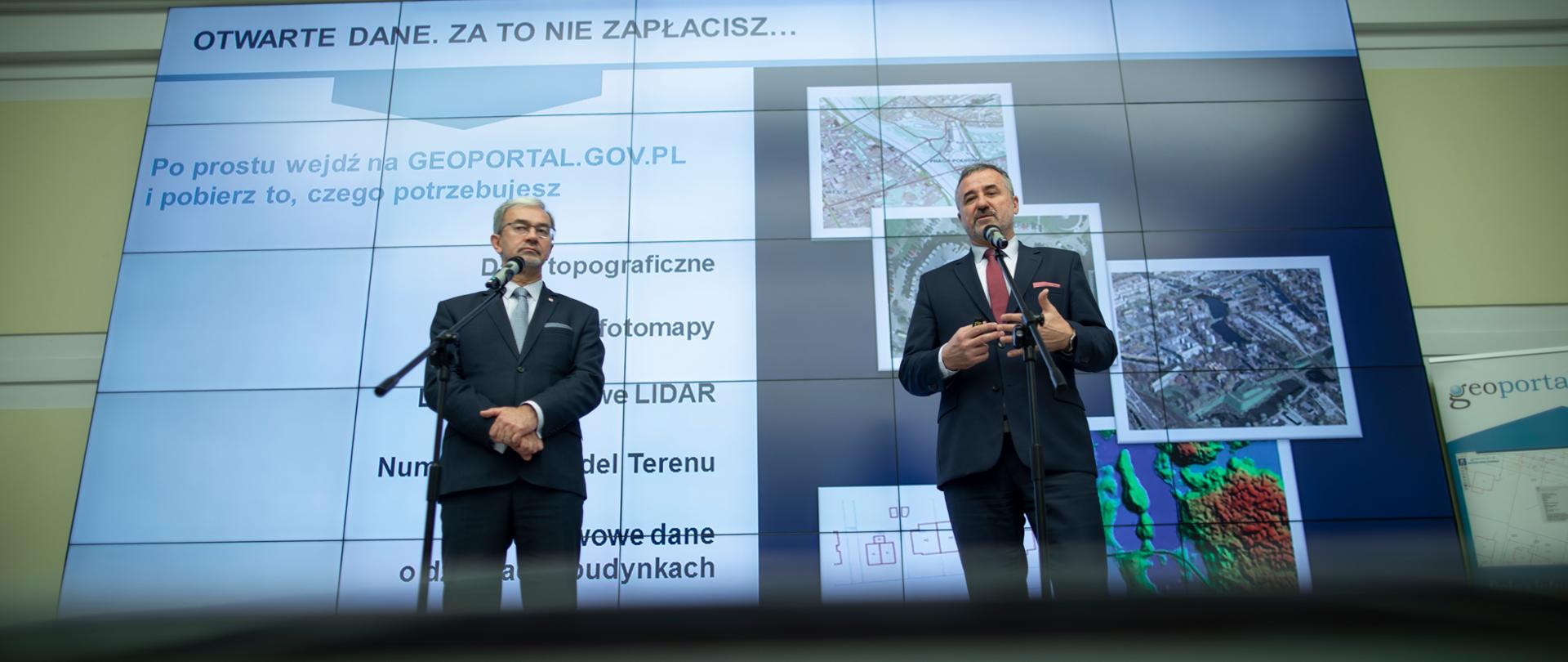 Na scenie przed mikrofonami stoją ministra Jerzy Kwieciński i Główny Geodeta Kraju Waldemar Izdebski. Za nimi na ekranie wyświetlona jest prezentacja.