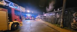 Na zdjęciu widać dwa samochody pożarnicze. Po prawej stronie zdjęcia nad budynkami unosi się dym. Jest pora nocna