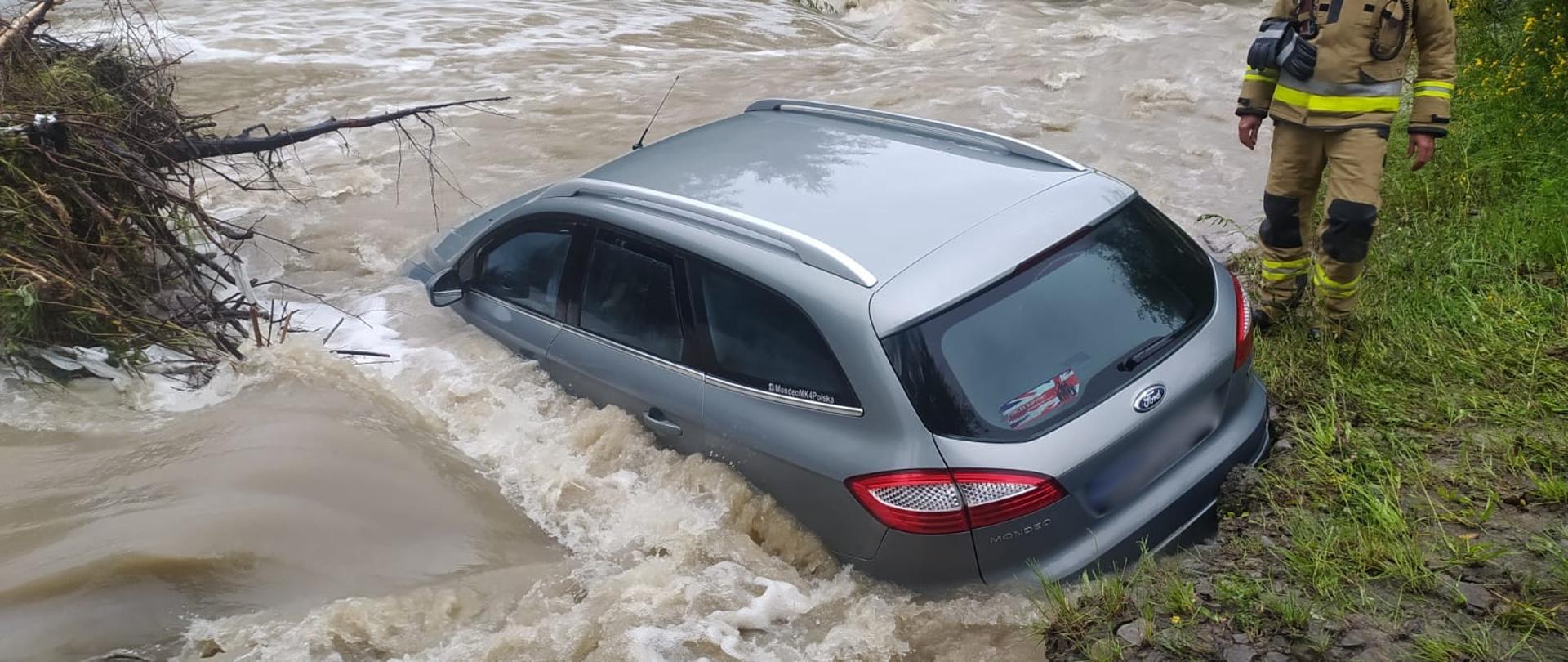 Samochód osobowy w rzece a obok strażak