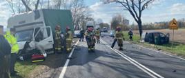 na zdjęciu droga oraz porozbijane elementy pojazdów jedne ciemny znajduje się na poboczu drogi w rowie drugi typu ciężarowego na drugim poboczu. Na środku drogi grupa strażaków za nimi samochody stojące w korku wokół drzewa oraz pole przed samochodem typu ciężarowego stoją dwie oosby.