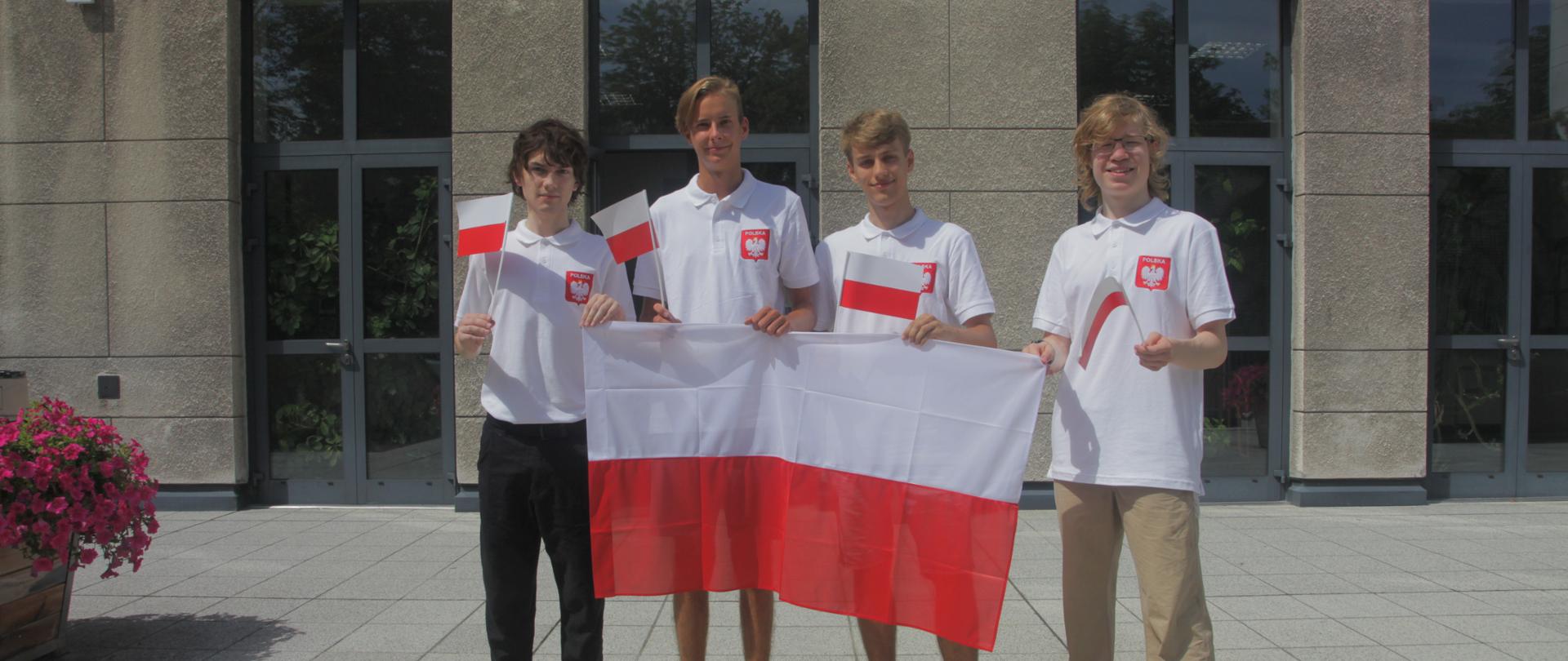 Zdjęcie przedstawia czterech uczestników 53. Miedzynarodowej Olimpiady Chemicznej. Tłem zdjęcia jest budynek Wydziału Chemii UW. Na zdjęciu chłopcy przed sobą trzymają biało czerwoną dużą flagę oraz w dłoniach małe flagi Polski. Ubrani w białe koszulki z godłem na piersi. 