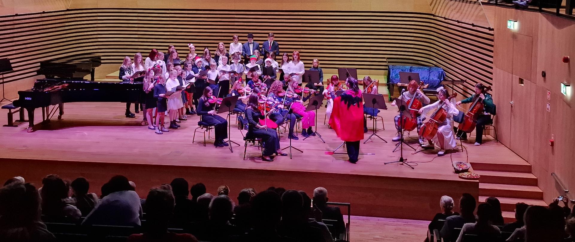 Grająca Orkiestra smyczkowa i śpiewający chór PSM I i II st. na scenie sali koncertowej z dyrygentką w stroju diabła