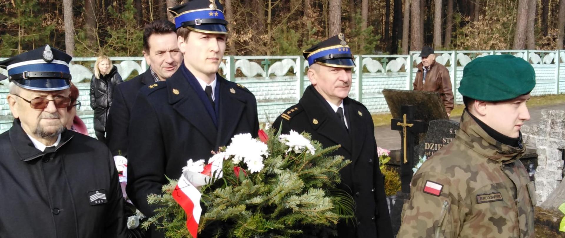 Trzech strażaków stoi na cmentarzu. Dwóch z nich to funkcjonariusze Państwowej Straży Pożarnej a jeden to druh OSP. Strażak stojący w środku trzyma w rękach wiązankę z biało czerwonych kwiatów. Przed nimi stoi żołnierz. Za strażakami widać mężczyznę i kobietę. W tle widać las.
