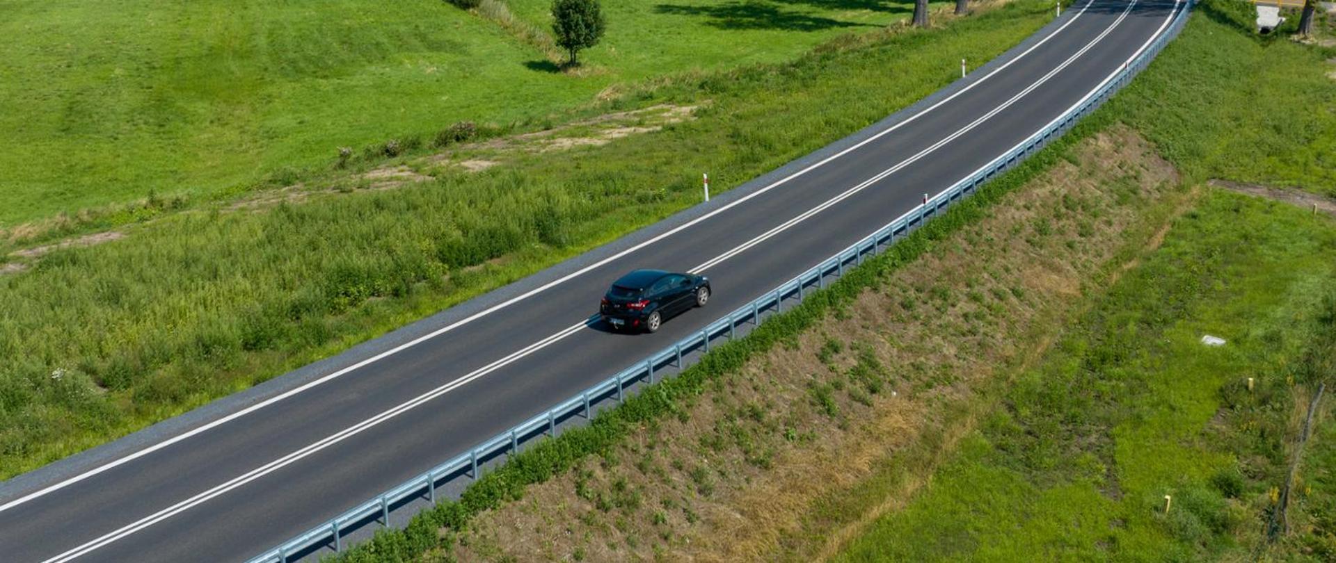 Widok z lotu ptaka na dwujezdniową drogę przebiegającą przez zielone tereny wiejskie. Po jezdni porusza się czarny samochód osobowy. Wzdłuż drogi znajdują się bariery ochronne, a wokół rozciągają się trawiaste pola i pojedyncze drzewa.