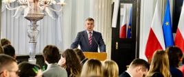 Na wypełnionej siedzącymi za stołami ludźmi sali za mównicą stoi minister Wieczorek i mówi do dwóch mikrofonów, obok niego elegancka szklano-złota lampa, z boku flagi Polski i UE.