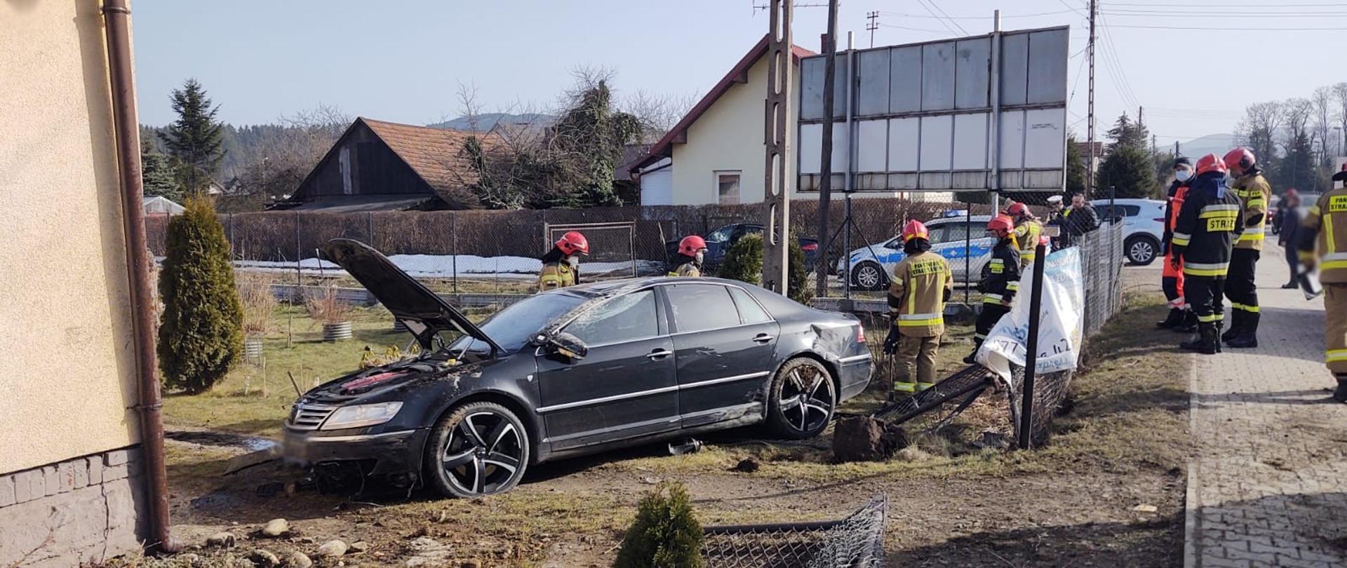 Samochód w ogrodzie obok domu oraz słupa betonowego. Wokół strażacy prowadzany działania. W tle inne budynki.