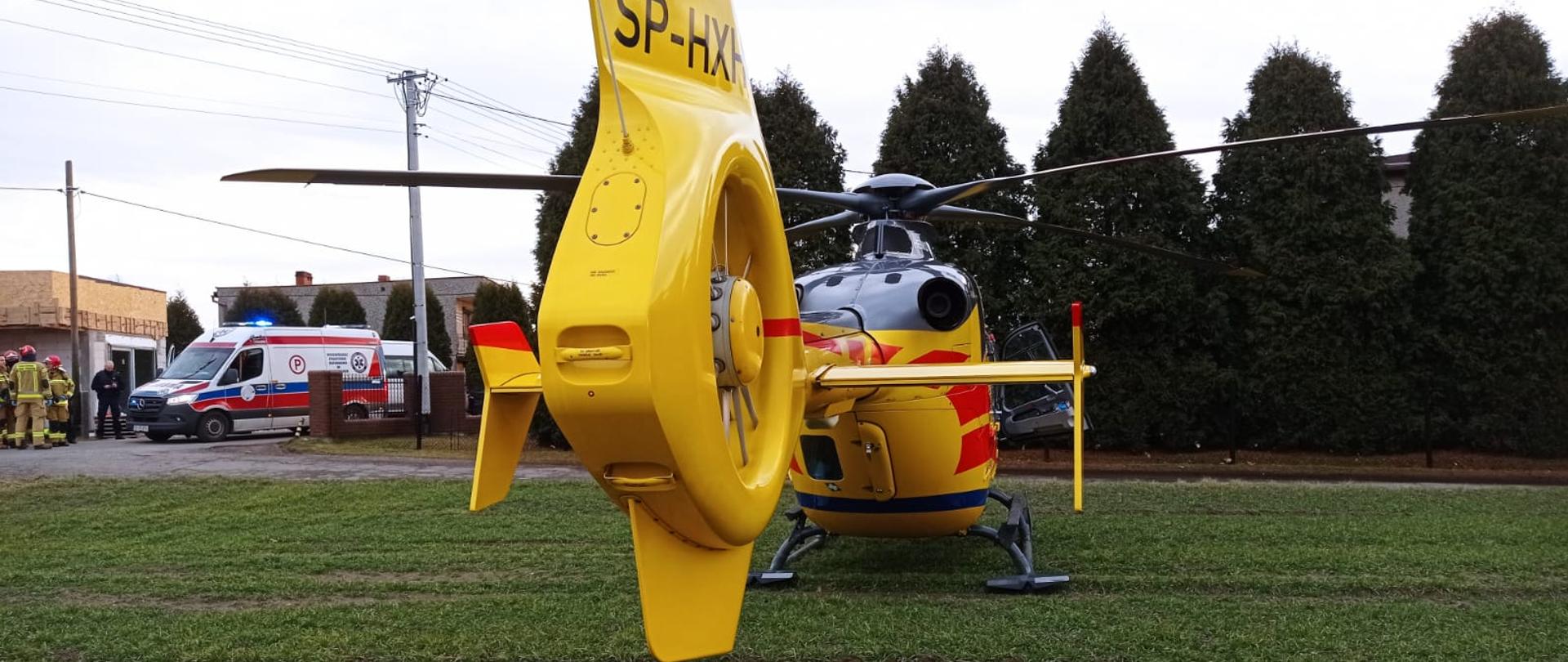 Śmigłowiec LPR stojący tyłem na trawie, w tle wysokie tuje, samochód pogotowia oraz strażacy.