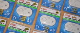 Kilka czujników tlenku węgla w opakowaniu. Urządzenia są podłużne, koloru białego, znajdują się na nich napisy. W zestawie trzy baterie "paluszki".