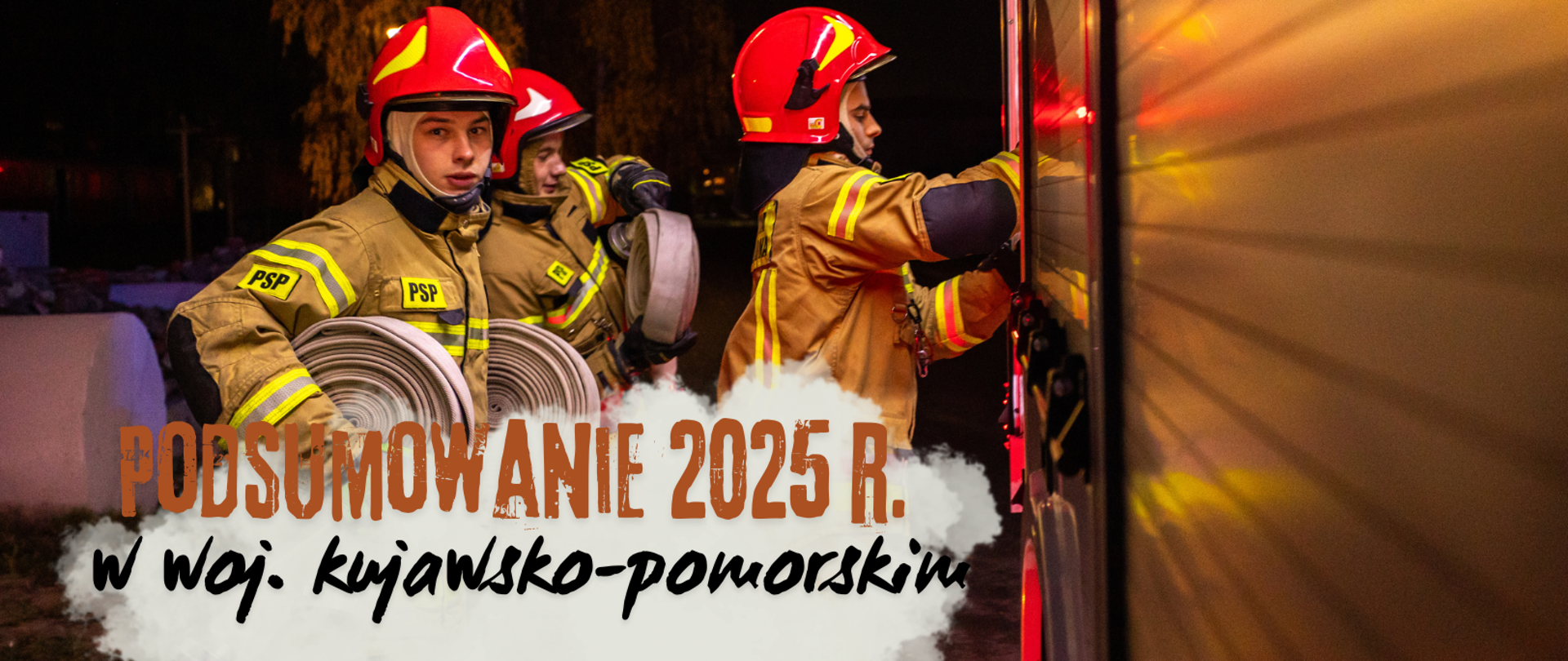 Na zdjęciu trzech strażaków w mundurach specjalnych, w hełmach na głowach, pierwszy trzyma wąż, inny zwrócony jest w stronę skrytki samochodowej. Z prawej strony widać bok samochodu.