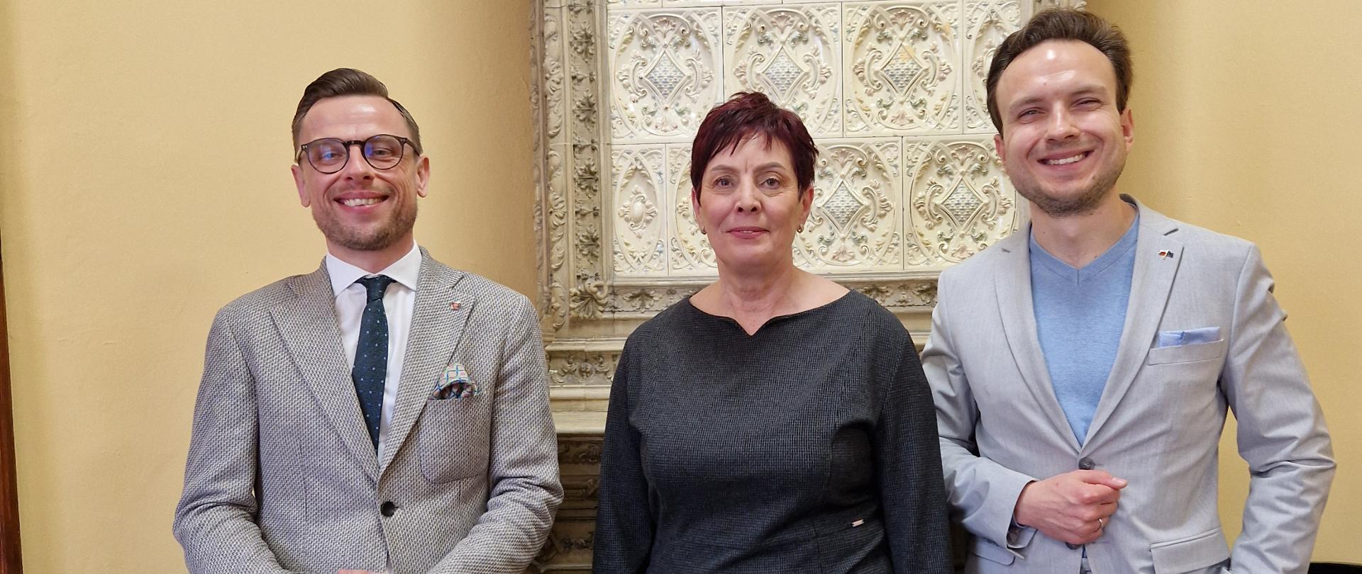 Fotografia prezentuje Wicewojewodę Dolnośląskiego p. dr Piotra S. Kozdrowickiego, Wojewódzkiego Inspektora Inspekcji Handlowej we Wrocławiu - p. Tomasza Reslera, Panią Dyrektor Delegatury Wojewódzkiego Inspektoratu Inspekcji Handlowej w Legnicy p. Annę Sapiełło-Renk. W tle piec kaflowy. 