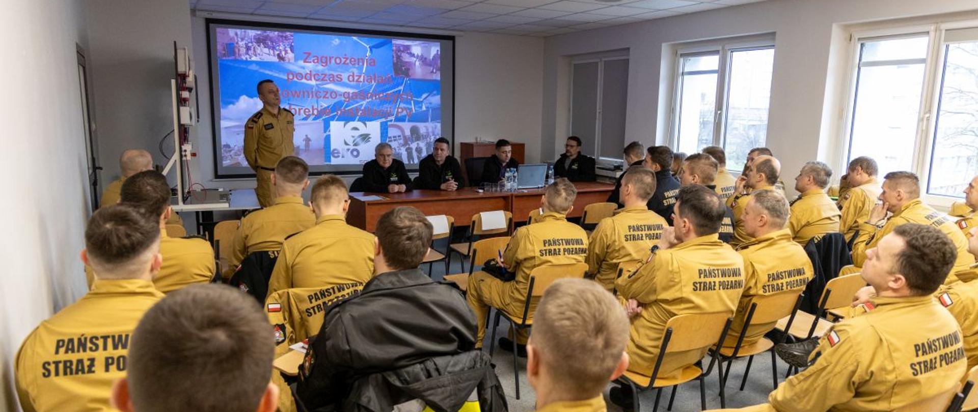Szkolenie doskonalące w zakresie zagrożeń podczas działań ratowniczych w obrębie paneli fotowoltaicznych. Strażacy Państwowej Straży Pożarnej w ubraniu służbowym siedzą na sali wykładowej Ośrodka Szkolenia Komendy Wojewódzkiej Państwowej Straży Pożarnej w Opolu słuchając przywitania dokonanego przez Zastępcę Opolskiego Komendanta Wojewódzkiego PSP w Opolu st. bryg. Arkadiusza Kuśmierskiego. Przed biurkiem siedzą przedstawiciele firmy szkolącej. Na ścianie wyświetlany jeden ze slajdów prezentacji.
