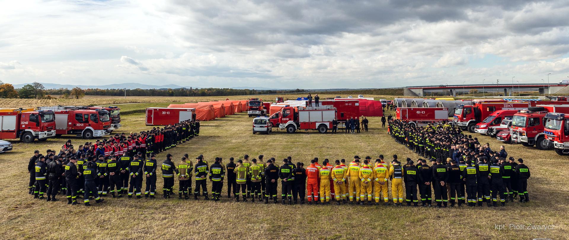 zdjęcie przedstawia stojących na łące ,w szeregach strażaków biorących udział w ćwiczeniach Opolskie 2021. Na dalszym planie samochody pożarnicze i namioty pneumatyczne. 