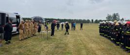 Zdjęcie przedstawia strażaków OSP ustawionych po prawej stronie w dwuszeregu. Ubrani są w ubranie koszarowe, pasy strażackie oraz hełmy. Strażacy stoją na boisku z trawą. Po lewej znajduje się samochód pożarniczy oraz kadra sędziowska i wójtowie gmin.