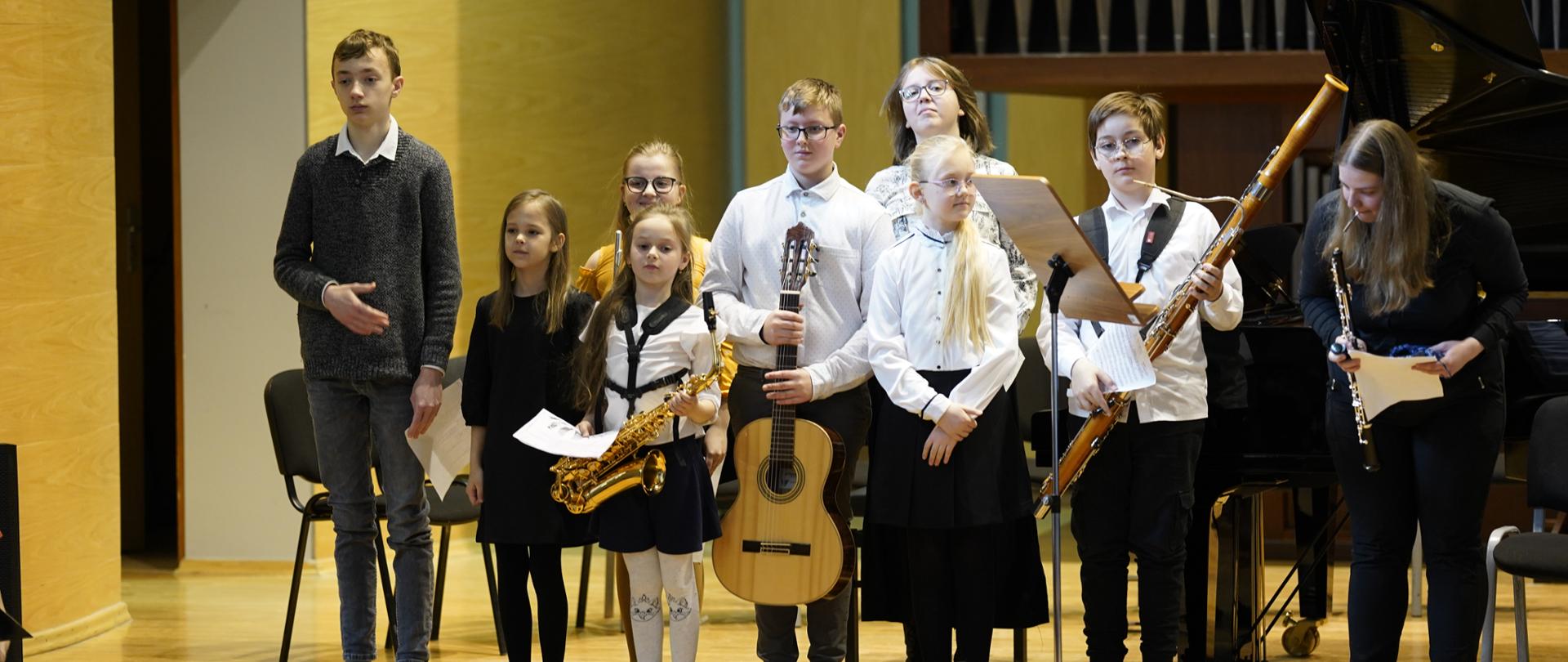 Uczniowie na scenie sali koncertowej w zdjęciu grupowym. Na pierwszym planie rozmazana widownia. Uczniowie stoją w jednym rzędzie trzymając swoje instrumenty muzyczne, przed nimi pulpit, za nimi fortepian, puste krzesła. W tle organy.