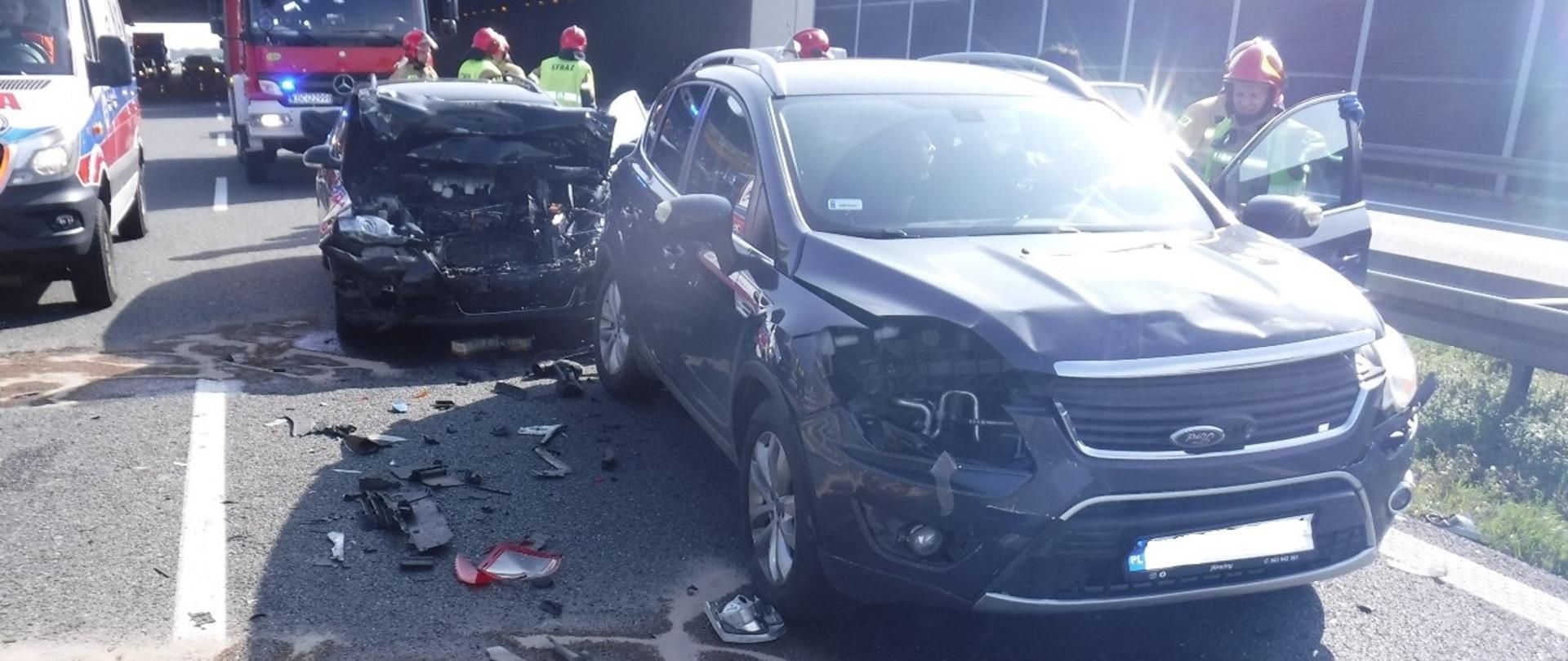 Zdjęcie przedstawia dwa rozbite somochody osobowe stojące na lewym pasie autostardy A4 w kierunku Krakowa. Na pierwszym planie stoi samochód marki Ford z rozbitymi przednimi reflektorami wgniecioną przednią maską oraz ułamanym przednim zderzakiem. Wokół samochodu leżą porozrzucane części samochodu, które oderwały się od niego. Za Fordem stoi kolejny zdeformowany pojazd z wgniecionym praktycznie całym przodem samochodu. Maska jest uniesiona do góry, a z pod samochodu wyciekają płyny eksploatacyjne na jezdnie, które są zasypane sorbentem. W lewym górnym rogu zdjęcia widać pojazd zespołu ratownictwa medycznego, tuż za nim stoi pojazd Straży Pożarnej z włączonymi światłami błyskowymi barwy niebieskiej. Na fotografi widać pięciu strażaków, którzy zabezpieczają pojazdy. Warunki atmosferyczne bardzo dobre słoneczna pogoda.