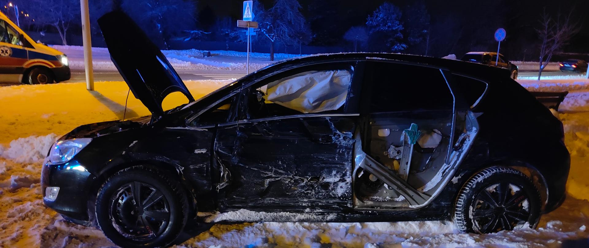 Na pierwszym planie stoi zaparkowany na poboczu drogi czarny samochód osobowy. Widać rozbity cały jego lewy bok. W tle stoi karetka pogotowia ratunkowego. Aura zimowa w porze nocnej