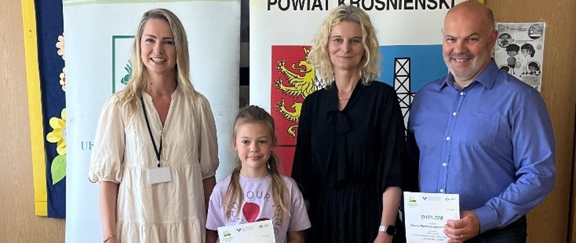 W pomieszczeniu na tle roll-upów stoją dwie kobiety i mężczyzna. Mężczyzna trzyma w rękach dyplom. Pomiędzy nimi stoi dziewczynka, która trzyma w rękach dyplom i torby z nagrodami.